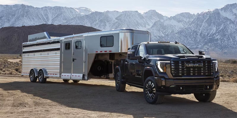 An all-new Black 2024 GMC Sierra 2500 HD hauling a large enclosed silver horse trailer parked amongst the mountains in the desert. - Boyd GM Oxford, NC