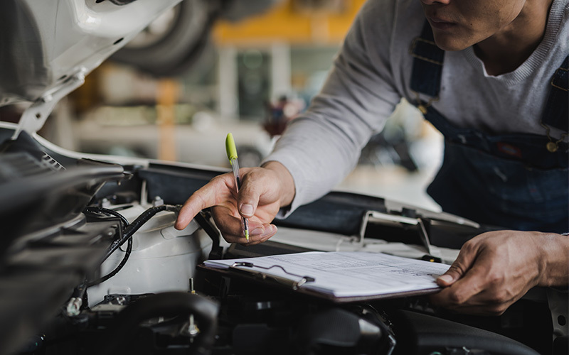 Technician inspecting a vehicle engine and recording maintenance details on a clipboard during a service inspection.
